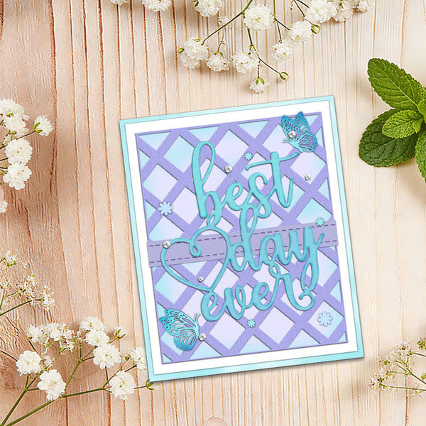 A handmade greeting card with the phrase 'best day ever' in turquoise script over a lavender and light blue lattice pattern, decorated with butterfly embellishments and small rhinestones, displayed on a light wood background with baby's breath flowers and mint leaves.