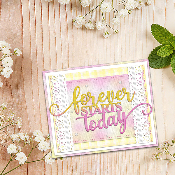 A handmade greeting card with the phrase 'forever STARTS today' in gold and pink script, layered over pastel pink, yellow, and white textured paper. The card is styled on a light wood background with white baby's breath flowers and mint leaves.