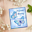 A handmade greeting card with blue and white butterfly illustrations and the words 'happiness blooming' in script, styled on a light wood background with baby's breath flowers and mint leaves.