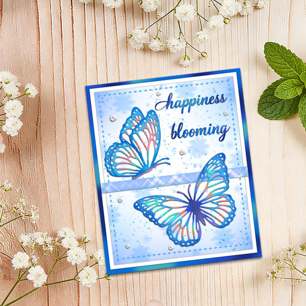 A handmade greeting card with blue and white butterfly illustrations and the words 'happiness blooming' in script, styled on a light wood background with baby's breath flowers and mint leaves.