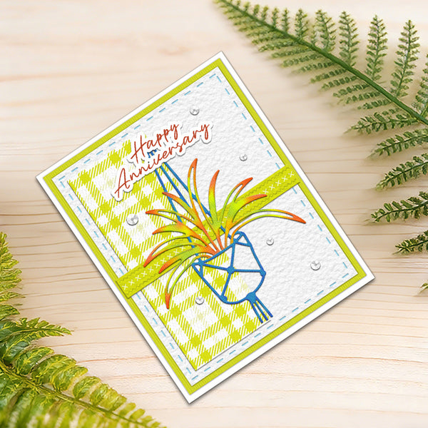 A handmade square greeting card with a 'Happy Anniversary' design, styled on a light wood surface with green fern leaves. The card features a white textured background, a lime green and white diagonal plaid border, a bright lime green horizontal stripe, and a blue geometric flower holding orange and yellow petals.