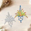 A metal die-cut craft template showing a plant in a pot design, with one side as a silver metal outline and the other side colored in with blue, yellow, and orange. The items are displayed on a light wood background with dried flowers and stones.