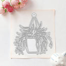 A metal die-cut craft stencil of a hanging plant in a pot, displayed on a square piece of white paper with pink roses in the top-left corner and a small white ceramic object in the bottom-right corner.