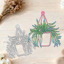 A metal die-cut craft template shaped like a hanging plant, shown in two states: one as a blank silver metal sheet and the other as a colorful filled template with green, blue, and pink layers. The items are displayed on a light wood background with dried flowers and small white flowers as props.