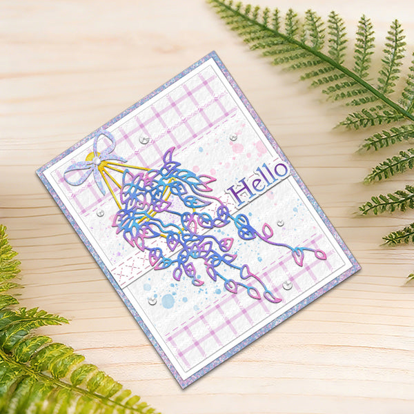 A handmade greeting card with a colorful abstract tree design, a lavender plaid pattern, and the word 'Hello' on a wooden surface, styled with green fern leaves.