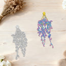 A decorative hanging ornament shown in two views on a light wood surface: a silver metal outline on the left and the finished iridescent multicolor ornament on the right with a yellow hanging loop. Dried flowers and stones are placed around the scene.
