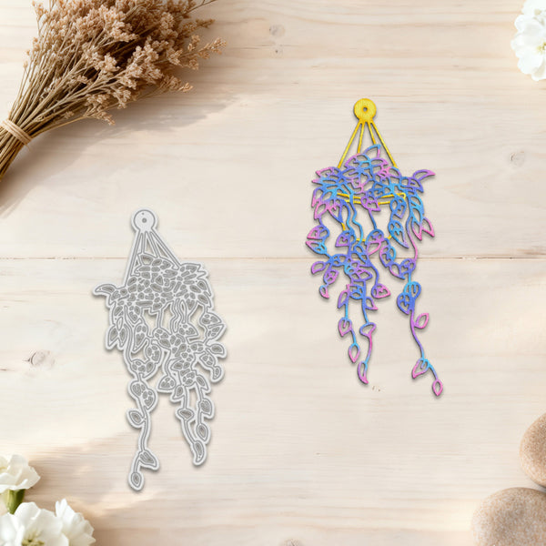 A decorative hanging ornament shown in two views on a light wood surface: a silver metal outline on the left and the finished iridescent multicolor ornament on the right with a yellow hanging loop. Dried flowers and stones are placed around the scene.