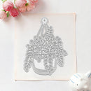 A hanging plant die-cut template or craft cutout shown on a square sheet of white cardstock, photographed on a light background with pink flowers in the top left and a small white ceramic object in the bottom right.