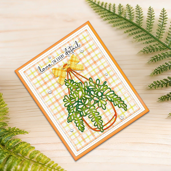 A square handmade greeting card with a yellow-orange border, featuring a green hanging plant illustration, a yellow bow, and the phrase 'love in even detail' on a light plaid background, styled on a light wood surface with green fern leaves.
