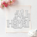 A metal die-cut craft stencil with the phrase 'Love is Home' in decorative lettering, displayed on a square sheet of craft foam with pink flowers and a small white pig figurine as props.