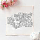 A floral and butterfly metal cutting die set displayed on a square piece of light pink paper, styled with pink roses in the top-left corner and a small white ceramic pig in the bottom-right corner.