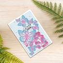 A handmade greeting card with layered pink and blue butterfly and floral die-cut designs on a light blue textured background, featuring the words 'Good luck' in black script. The card is displayed on a light wood surface with green fern leaves in the corners.