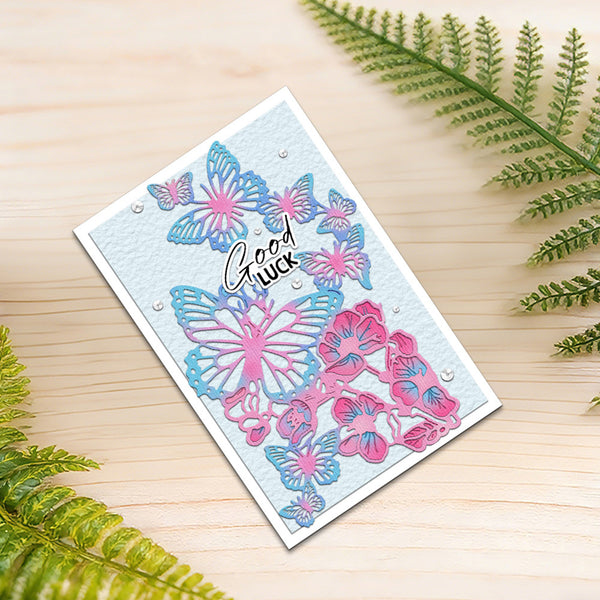 A handmade greeting card with layered pink and blue butterfly and floral die-cut designs on a light blue textured background, featuring the words 'Good luck' in black script. The card is displayed on a light wood surface with green fern leaves in the corners.