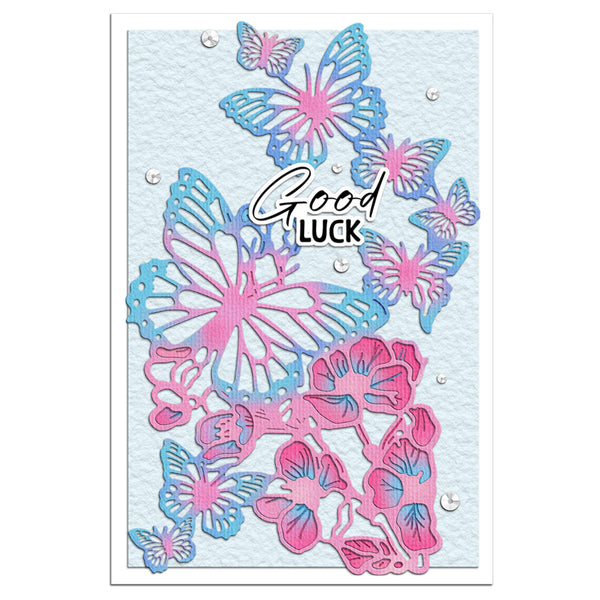 A square greeting card featuring layered die-cut butterflies and flowers in pink and blue on a light blue textured background, with the words 'Good Luck' in white script and small clear rhinestone accents.