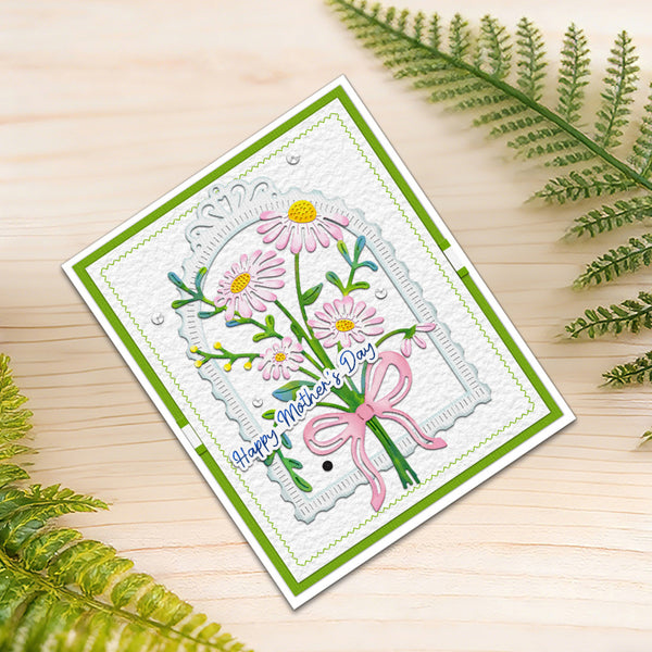 A handmade Mother's Day greeting card with a floral design, placed on a light wood surface with green fern leaves around it. The card features pink daisies, green stems, a pink bow, and the text 'Happy Mother's Day'.