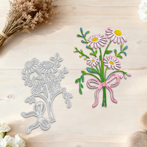 A floral die-cut craft design shown on a light wood surface, featuring a bouquet of daisies with pink petals, yellow centers, green stems, and a pink ribbon. The left side also shows the corresponding silver metal die cutout shape.