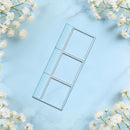 A rectangular metal stamp or stencil with three elongated square cutouts, shown on a light blue marble background with white baby's breath flowers around the edges.