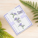 A handmade square greeting card with a white embossed background, lavender borders, floral sprig illustrations, and a 'Happy Birthday' message, styled on a light wood surface with green fern leaves.