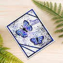 A square handmade greeting card with two blue and purple butterflies, black ink details, and a 'Happy Birthday' message, displayed on a light wood surface with green fern leaves.