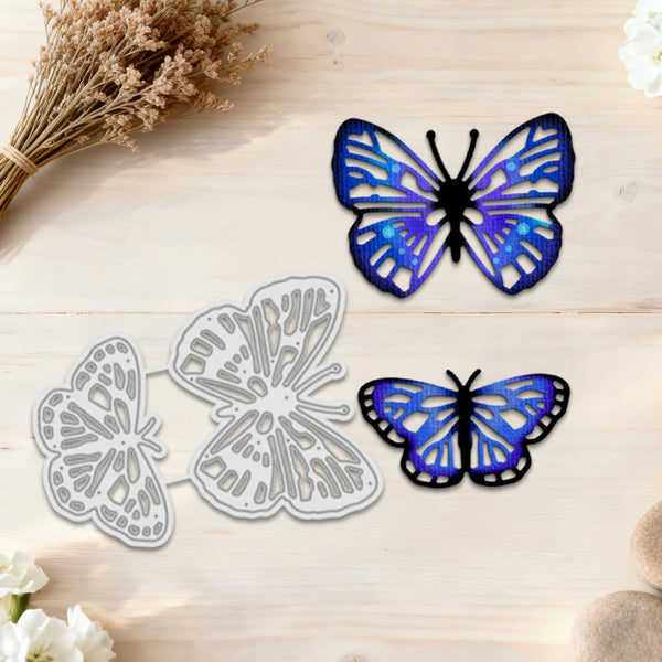 Three butterfly-shaped craft dies or embellishments arranged on a light wood surface: two silver metal outlines and one blue-and-purple butterfly piece with black outlines. Dried flowers and small stones decorate the corners.