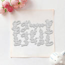 A flat sheet of beige cardstock or paper with a large outlined decorative script design in the center, styled on a white surface with pink flowers in the top left and a small white ceramic object in the bottom right.
