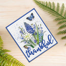 A handmade square greeting card with a blue border and the word 'thankful' in blue script, decorated with blue flowers, green leaves, a butterfly, and a small bee on a light wood surface with fern leaves around it.