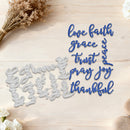 A flat-lay of a metal die-cut craft cutting mat featuring the words 'love faith grace trust pray joy thankful' in blue script, shown next to the silver metal cut shapes on a light wood background with dried flowers and white blossoms.
