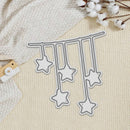A metal die-cut craft embellishment featuring five hanging star shapes on a string, displayed on a textured cream fabric background with cotton stems and a wooden camera prop.