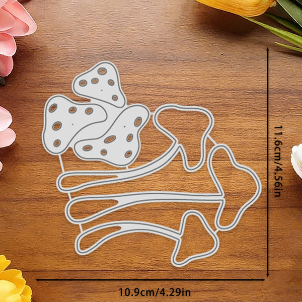 A metal die-cut craft template shaped like mushrooms and stems, shown on a wooden surface with printed dimension lines indicating 11.6 cm by 10.