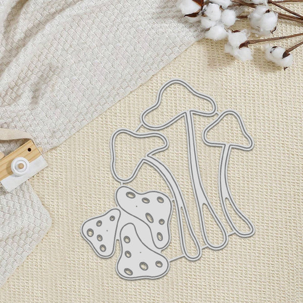A mushroom-shaped metal cutting die set with a silver-colored outline, displayed on a textured cream fabric background alongside a waffle-knit textile, cotton stems, and a small wooden-and-white camera accessory.