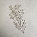 A metal floral die-cut stamp or embosser featuring a lily-of-the-valley design, shown on a textured off-white paper background.