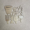 A set of metal garden tool and flower cutters or molds arranged on a textured off-white background. The set includes a flower pot, a fish-shaped piece, leafy vines, a trowel, shears, a rake, and a watering can.