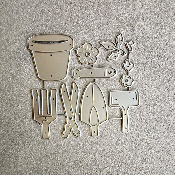 A set of metal garden tool and flower cutters or molds arranged on a textured off-white background. The set includes a flower pot, a fish-shaped piece, leafy vines, a trowel, shears, a rake, and a watering can.