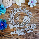 A silver metal die-cut craft embellishment featuring a bird and decorative scrollwork, displayed on a rustic wooden surface with decorative flowers and a candle partially visible.