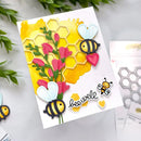 A handmade greeting card with a yellow honeycomb and watercolor background, pink flowers, two die-cut bees, hearts, and the word 'bee well' in script. The card is displayed on a light surface with some green leaves and crafting tools in the background.