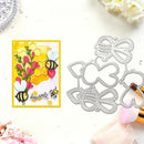 A flat-lay product photo showing a handmade greeting card with a yellow border, pink flowers, honeycomb pattern, and two cartoon bees, alongside two metal die-cutting templates shaped like bees and flowers. The card and dies are arranged on a white surface with decorative flowers and makeup brushes around them.