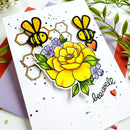 A handmade greeting card featuring a large yellow flower, two bees, purple blossoms, and the word 'be well' in black script, styled on top of colorful envelopes with green leaves in the background.