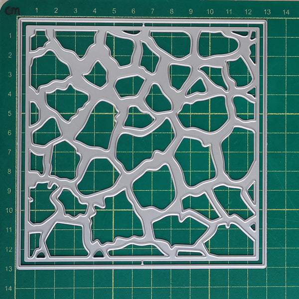 A square metal stencil with an organic, web-like cutout pattern, displayed flat on a green cutting mat with grid lines.