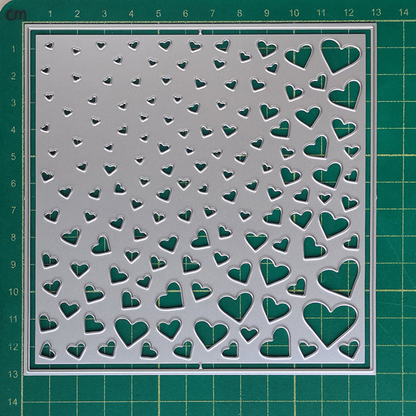 A square metal craft stencil with a pattern of small and large heart cutouts, displayed on a green cutting mat.