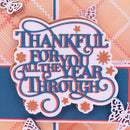 A close-up of a decorative layered paper cutout with the phrase “THANKFUL FOR YOU ALL THE YEAR THROUGH” in blue script with orange-red outlines, on a patterned scrapbook background with butterflies and stars.
