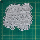 A silver metal die-cut craft stencil with Cyrillic lettering and floral motifs, displayed on a green cutting mat with grid lines and measurements.