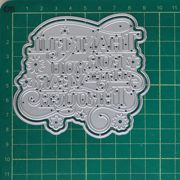 A silver metal die-cut craft stencil with Cyrillic lettering and floral motifs, displayed on a green cutting mat with grid lines and measurements.
