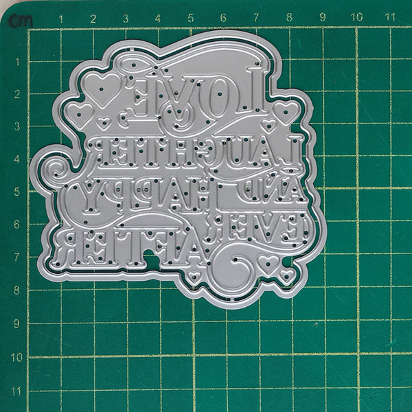 A silver metal die-cut craft embellishment with the words 'LOVE LAUGH AND HAPPY EVER AFTER' in decorative lettering, shown on a green cutting mat with grid lines.