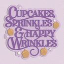 A decorative sign or plaque with the phrase “Cupcakes Sprinkles & Happy Wrinkles” in purple script, decorated with small tan cupcakes and white stars on a pale lavender background.