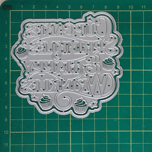 A silver metal cutting die with a decorative 'Happy Cupcakes, Pankakes, Weekakes' design, displayed on a green cutting mat with grid lines.