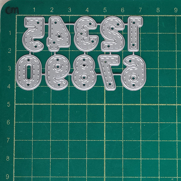 A set of metal letter cutters in the shape of stylized numbers and letters, arranged on a green cutting mat with grid lines.