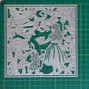 A square metal cutting die with an intricate Alice in Wonderland scene, featuring Alice, the White Rabbit, a mushroom, a clock, and a tree, displayed on a green cutting mat.