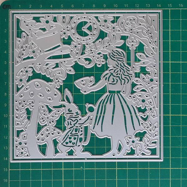A square metal cutting die with an intricate Alice in Wonderland scene, featuring Alice, the White Rabbit, a mushroom, a clock, and a tree, displayed on a green cutting mat.