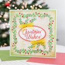 A square Christmas greeting card with a green holly and red berry border, yellow bows, and a central white oval reading “Christmas Wishes,” photographed on a tabletop with a blurred Christmas tree in the background.