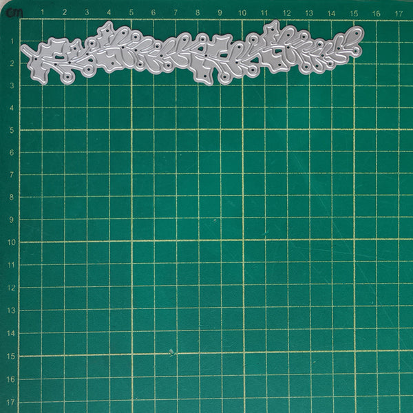 A silver metal die-cut floral border with leaf and vine patterns, displayed on a green cutting mat with centimeter grid lines.